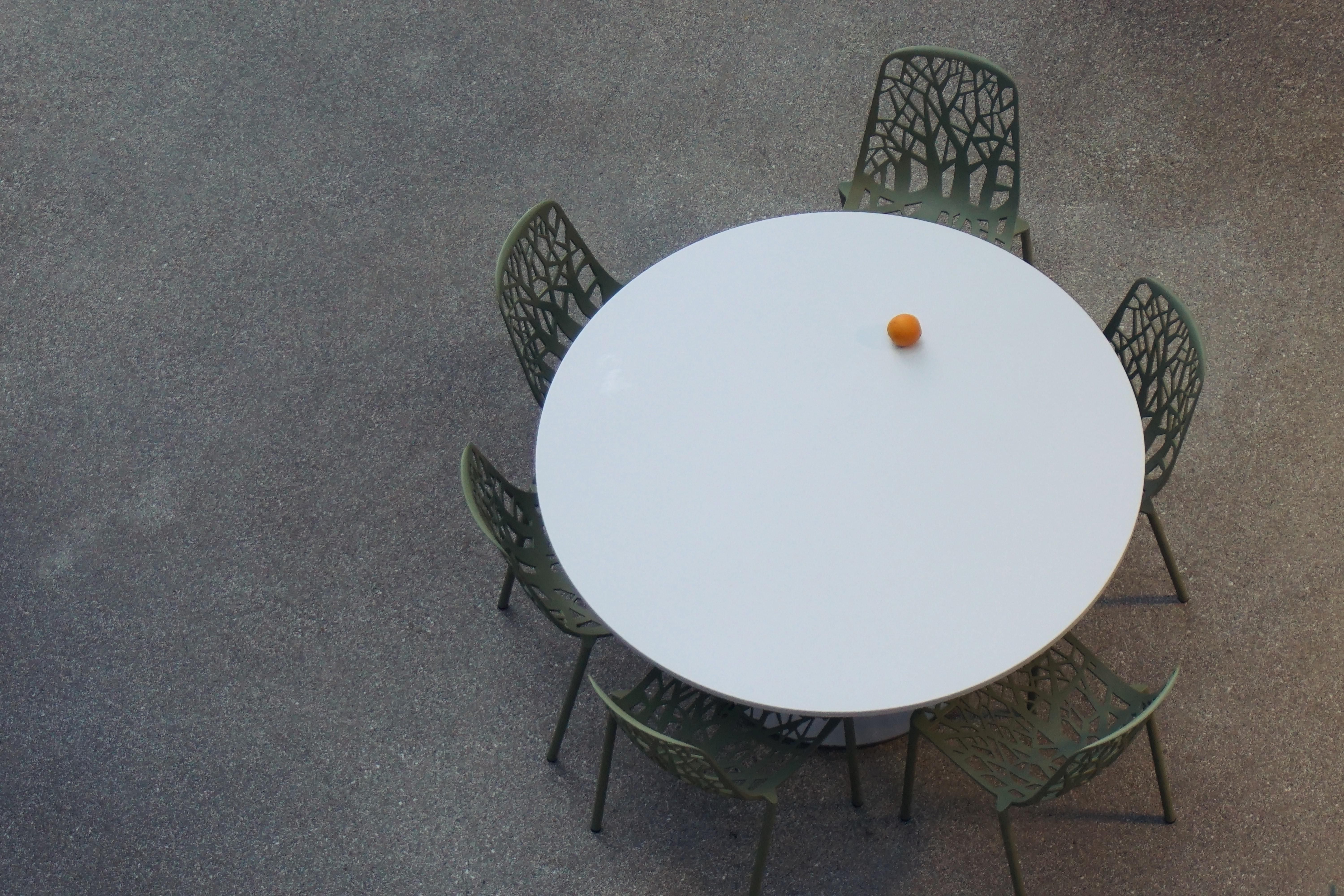 A photo of a circular table with six chairs. There is one lone orange on the table.
