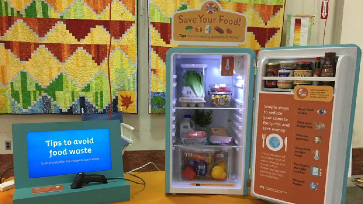 A photo of the "Save Your Food!" tabletop exhibit, featuring a mini fridge stocked with food and a grocery scanner.