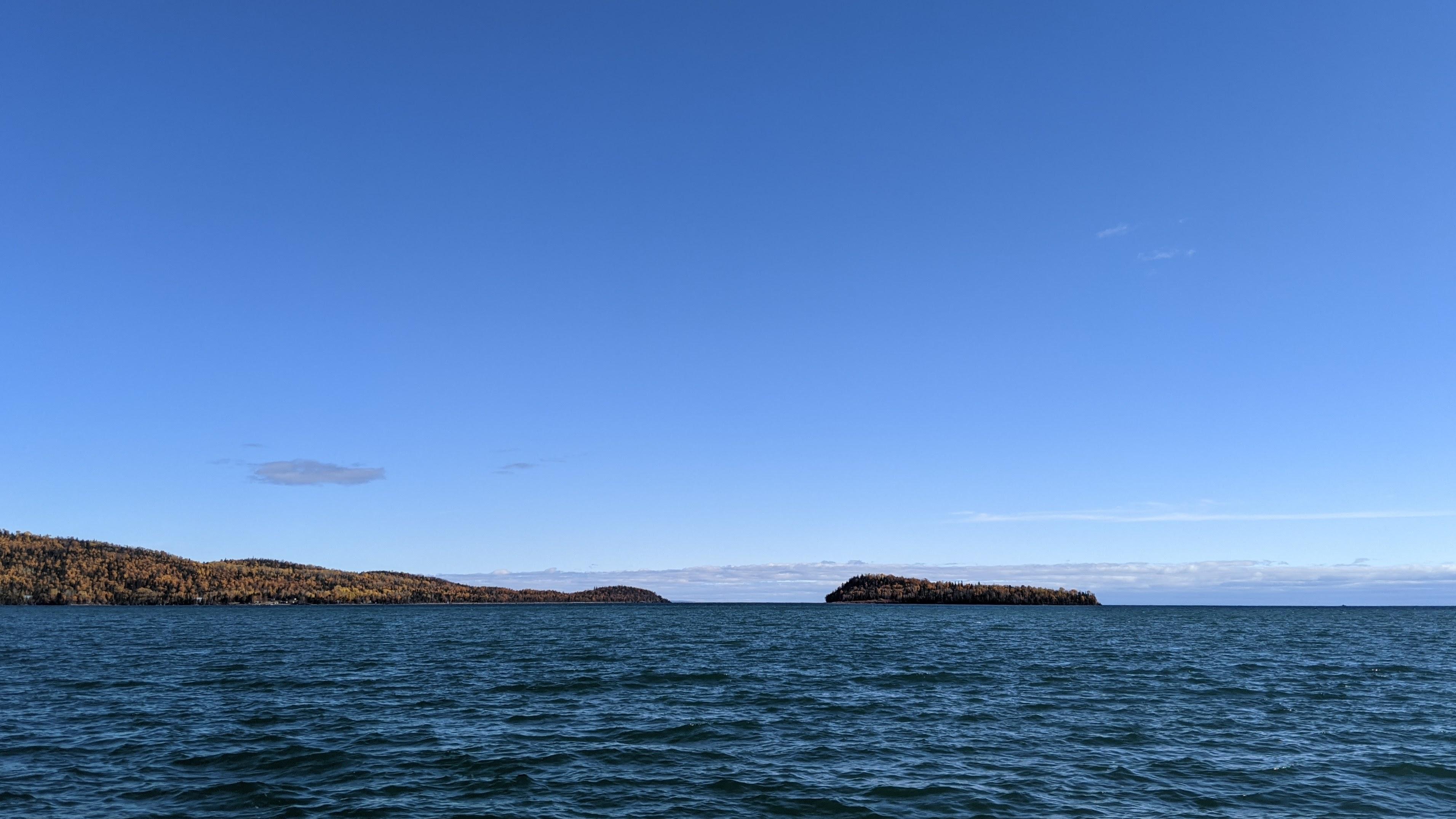 A photograph of Lake Superior with a forested peninsula at left nearby a small island, under a blue sky.