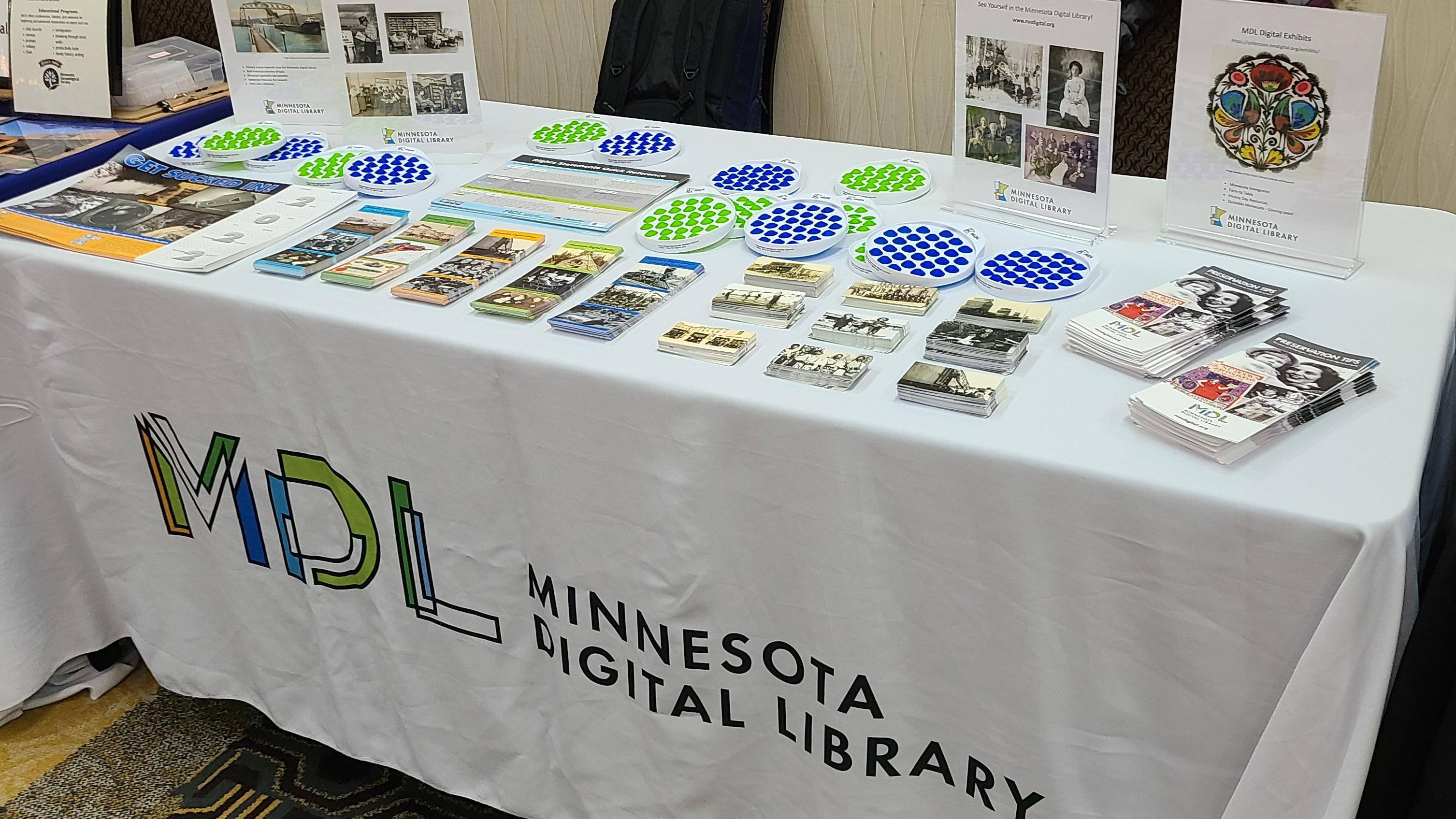 A table covered in a white tablecloth with the MDL logo on the front. The top  of the table has MDL promotional print materials and branded blue and green pop-it fidget toys