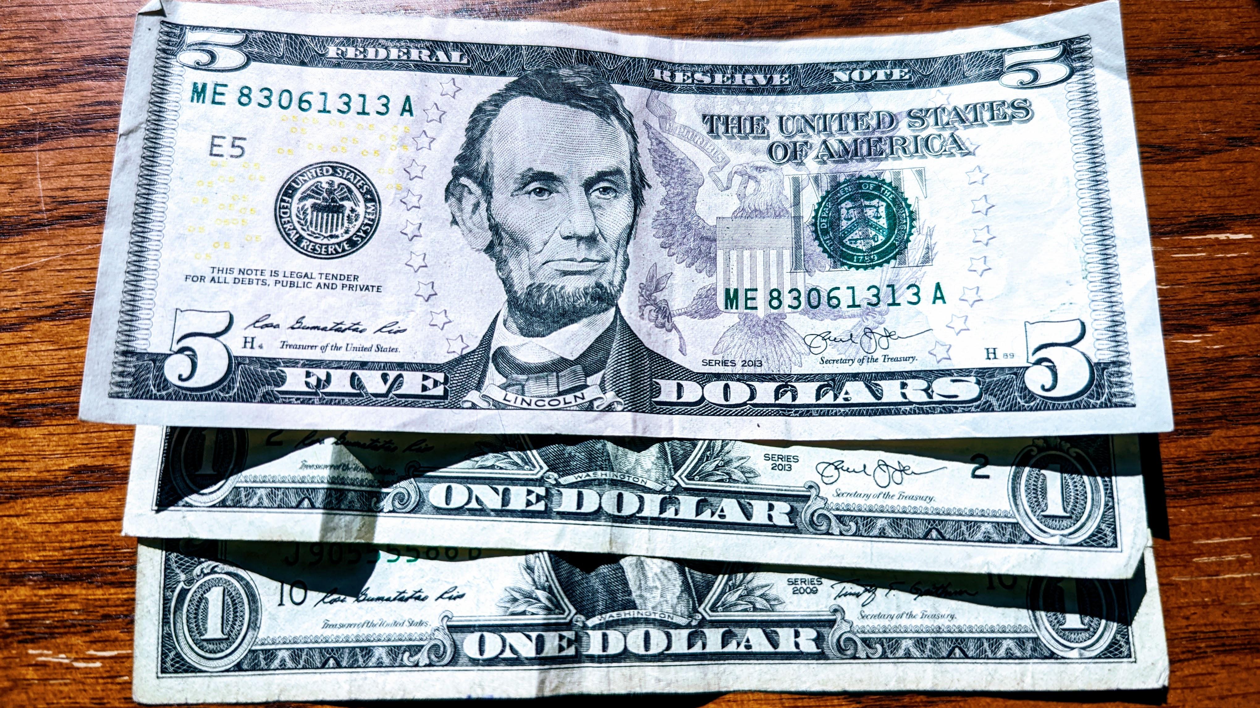 A photograph of a five-dollar bill atop two one-dollar bills.