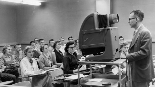 Man with a large overhead projector standing in front of a class of college students