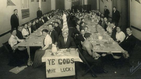 Group of male and female college sophmores seated at long tables with one man in a tall funny hat in the foreground