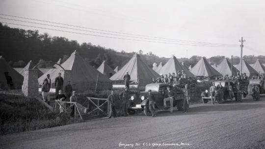 Row of white triangular tents lining a dirt road, with men and trucks lined up in front