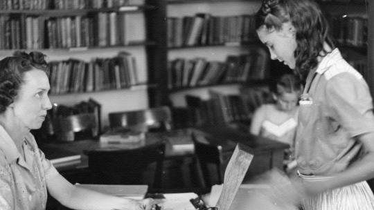 Librarian helps a student while seated at a desk in a library with books and papers