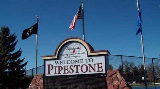 White outdoor sign with red and black letters reading Welcome to Pipestone. Sign is in front of a blue sky and three flags, one for POW/MIA, the United States flag, and the Minnesota State Flag