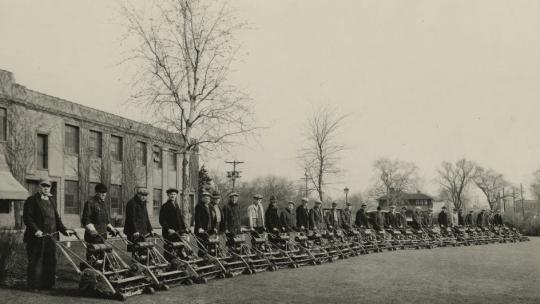 Men with Toro lawnmowers lined up in an arc in front of a building with trees in the background