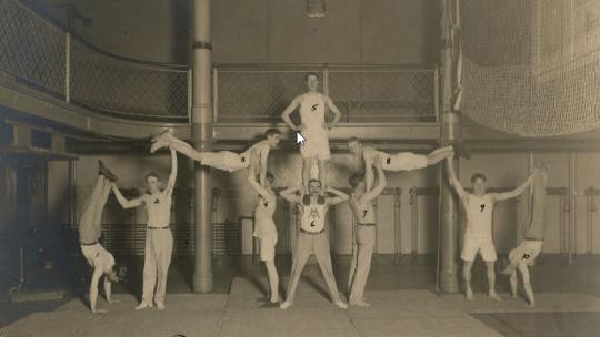 Eight men in exercise clothing balancing to form a gymnastic pyramid in a gymnasium