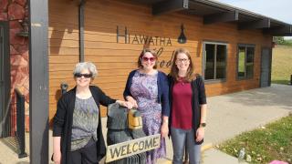 Three women standing outside in front of a long, low, wood-sided building named the Hiawatha Lodge