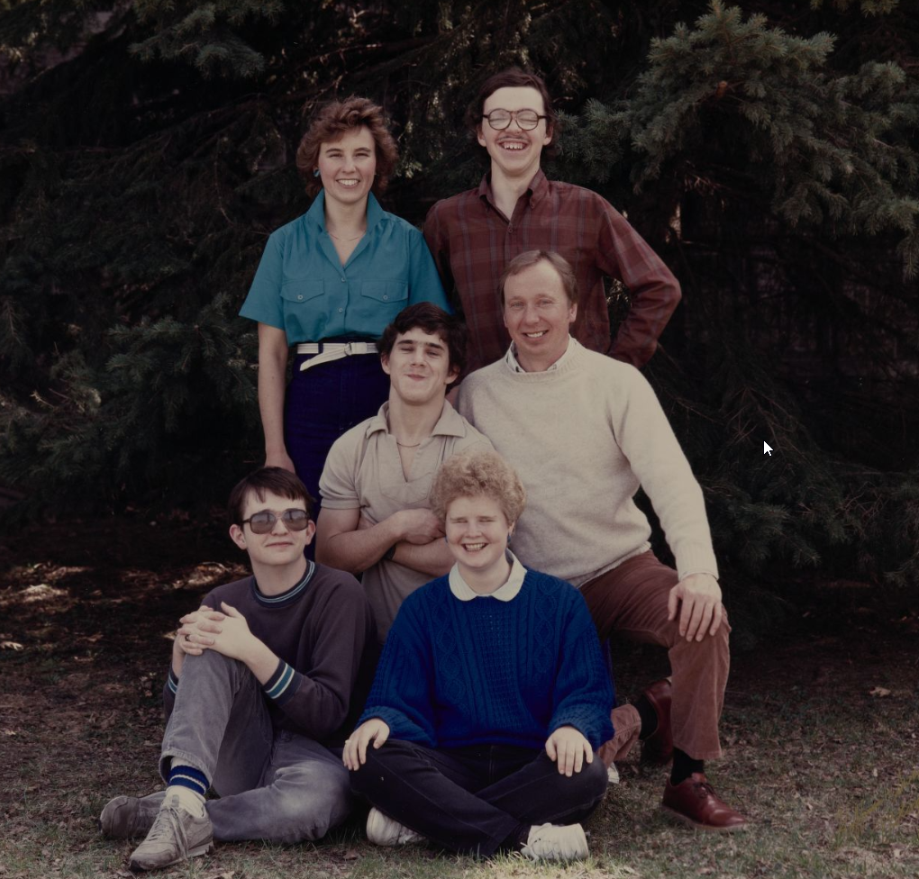 Six people, two standing, four seated, smiling towards a camera, posed in front of some trees outside during mild weather