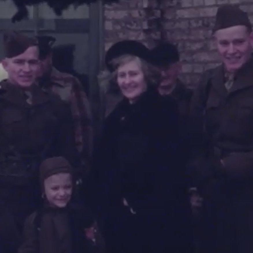 Two young men in military uniform standing with a woman and a young boy, both in hats and coats, in front of a brick wall