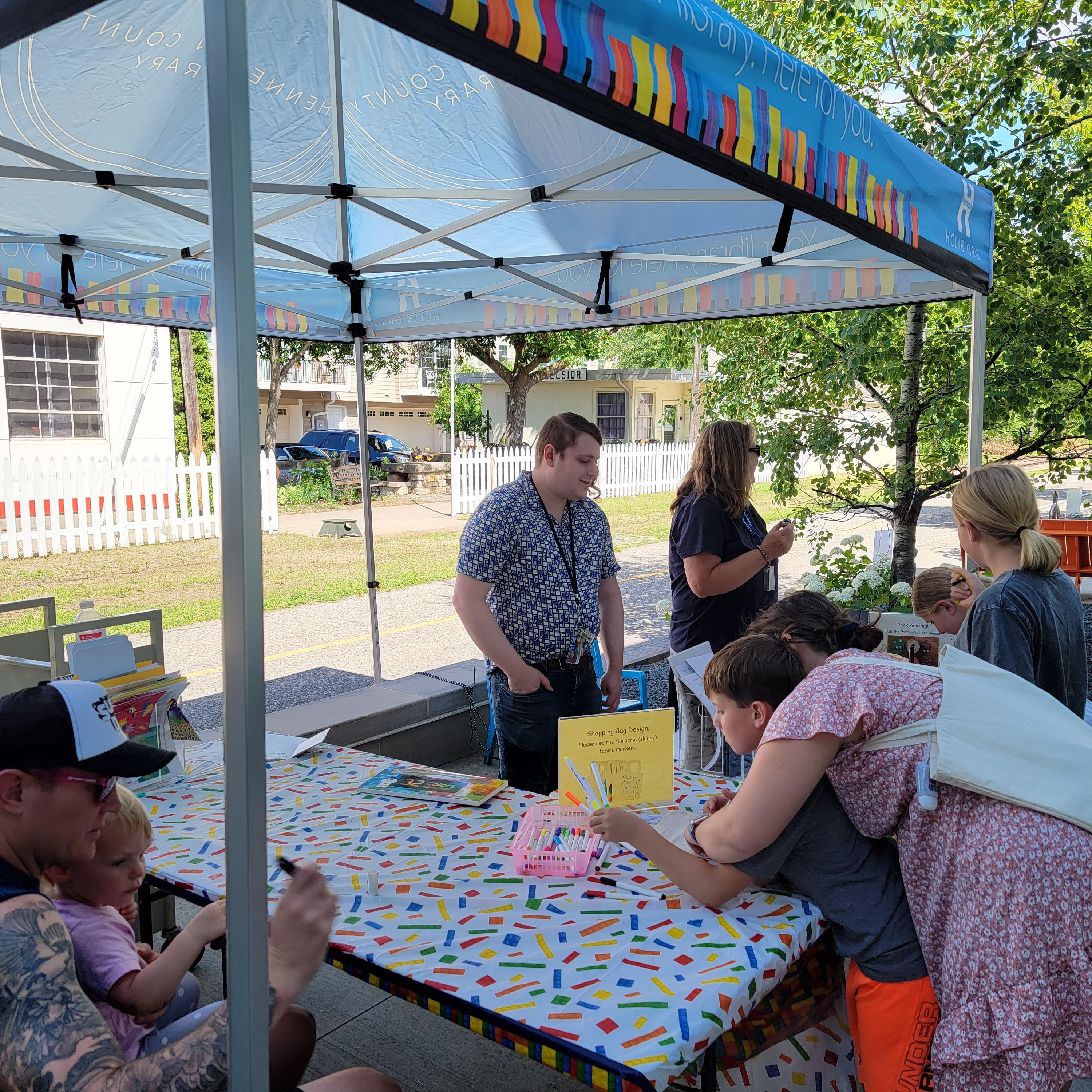 A photo of a tent outside of the Excelsior Library, with library staff supervising kids and adults as they work on crafts.