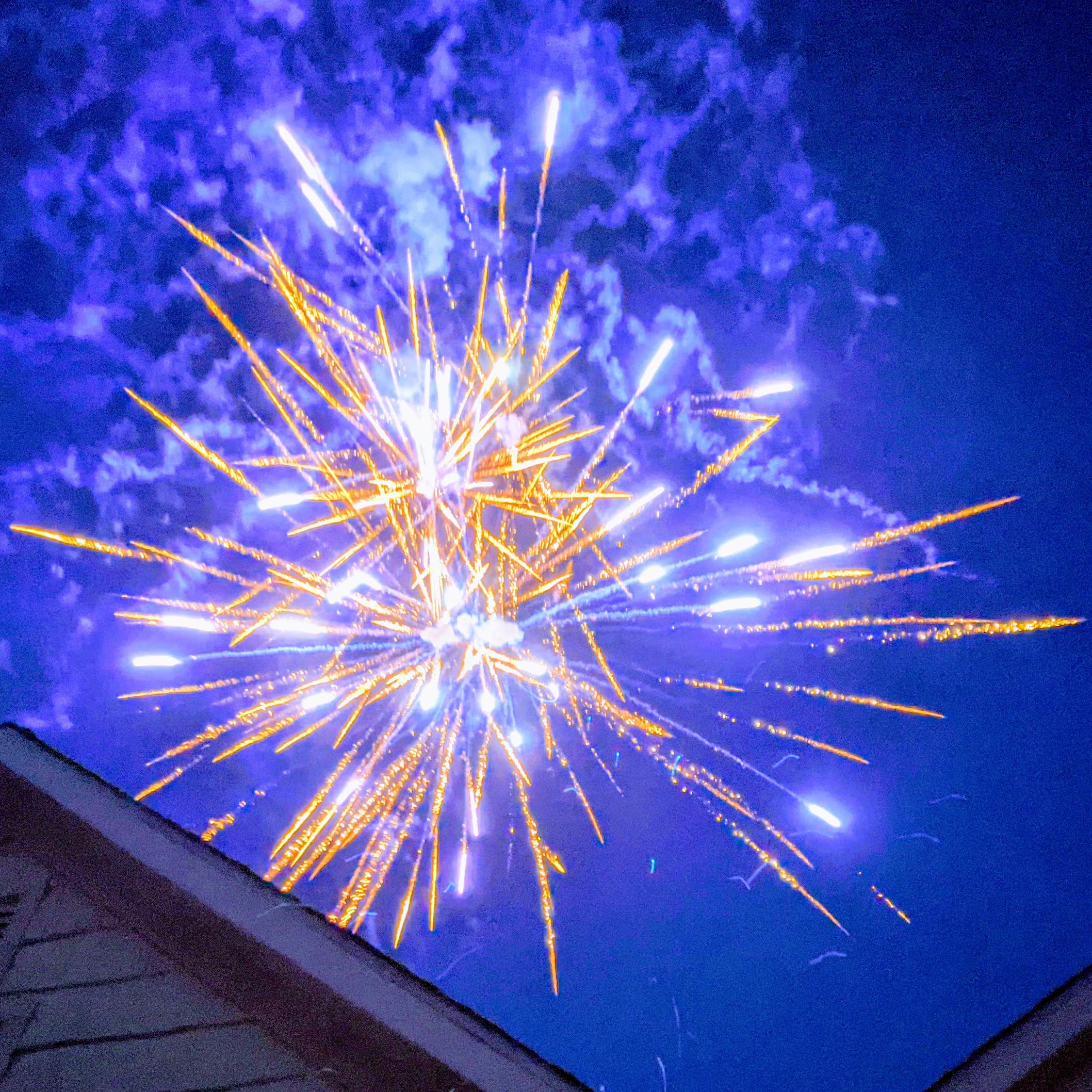White and yellow fireworks burst in a royal blue night sky above the outline of an American home.