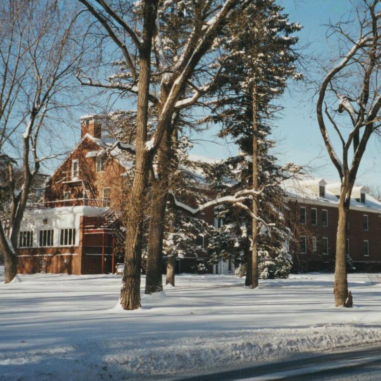 Multi-story brick building partially obscured by trees in the foreground. There is a light coating of snow on the ground and building roof.