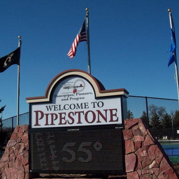 White outdoor sign with red and black letters reading Welcome to Pipestone. Sign is in front of a blue sky and three flags, one for POW/MIA, the United States flag, and the Minnesota State Flag