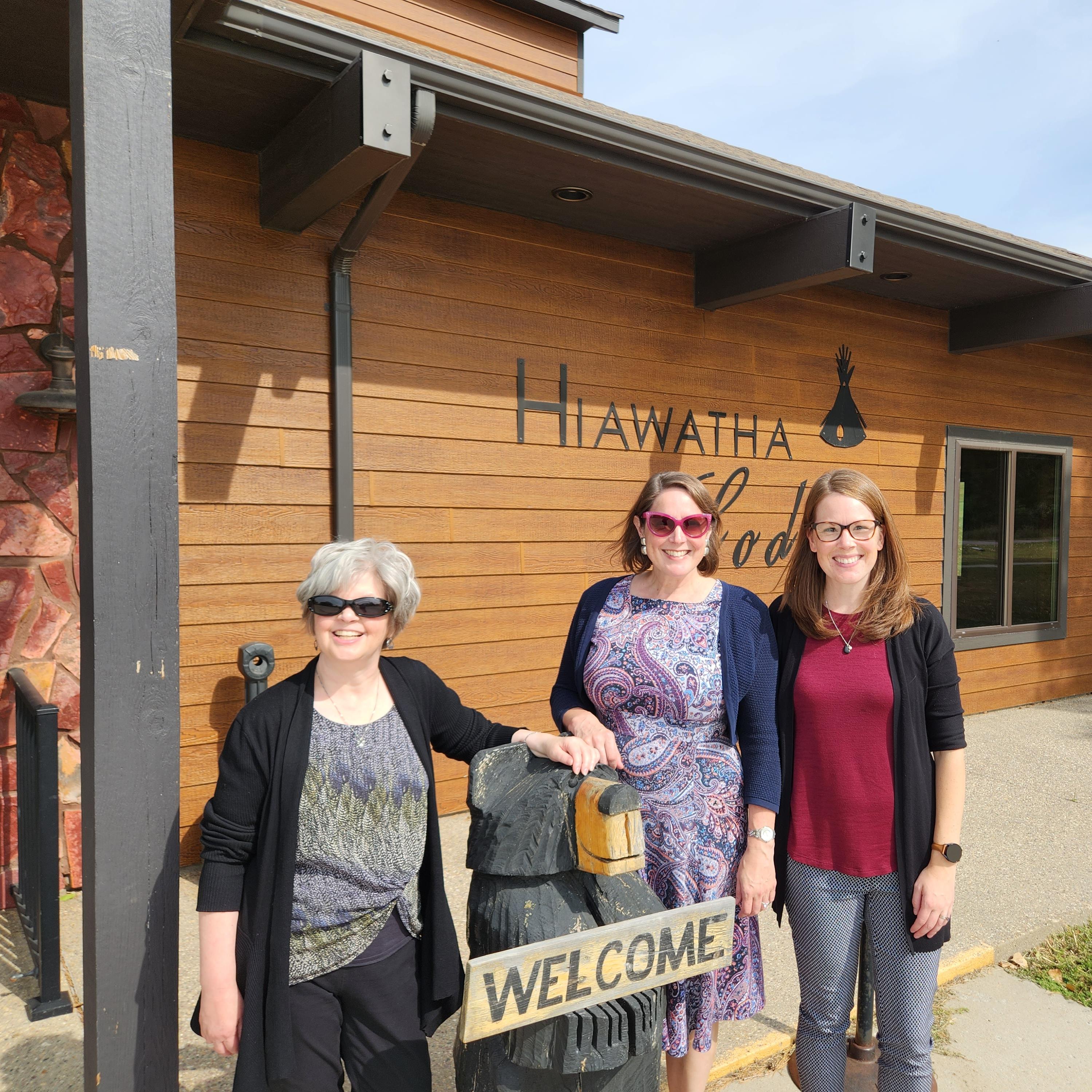 Three women standing outside in front of a long, low, wood-sided building named the Hiawatha Lodge
