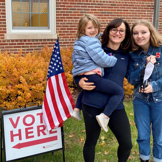 A photograph of Melanie Hazelip and her family next to an American flag and "vote here" sign.